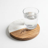 A round wooden and white marble coasters with a dark engraved mountain design, shown with a clear glass of water on a white background.