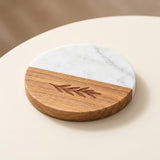 A round half-and-half serving board or trivet with a white marble top half and a natural wood bottom half featuring an engraved leaf design, shown on a light beige surface.