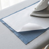 A close-up of a white fabric sheet or towel with blue edges laid on a light gray ironing board, with a white and gray iron resting on top.