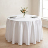 A round white tablecloth draped over a round table in a bright room, styled with two place settings and a small floral centerpiece in a vase.