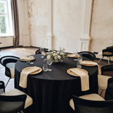 A round black tablecloth-set dining table in a rustic room, surrounded by black and cream chairs with beige napkins, plates, wine glasses, and a white floral centerpiece.