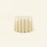 A round tablecloth in a light cream or ivory color, draped elegantly over a circular table against a solid, very light warm background.