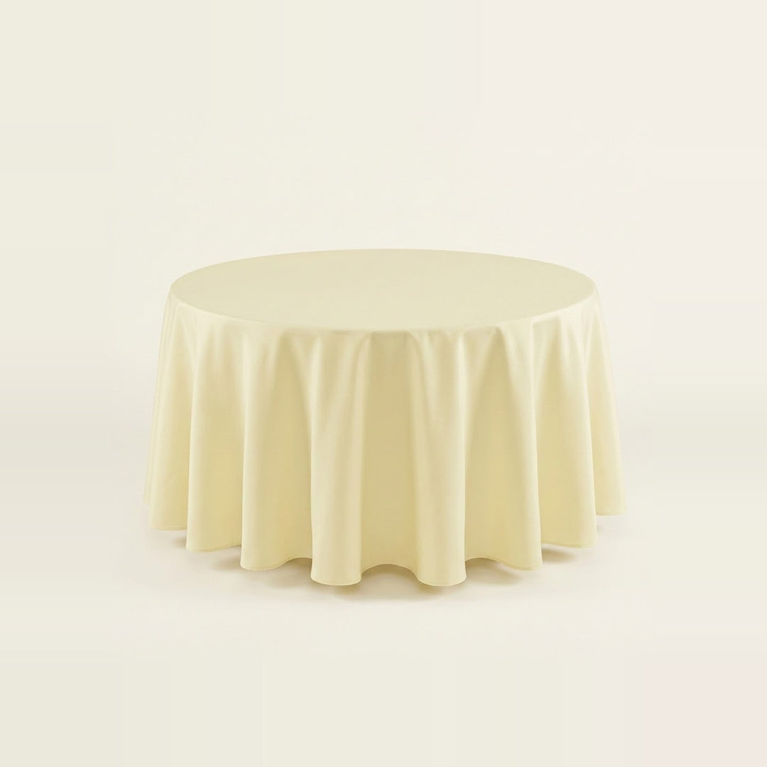 A round tablecloth in a soft cream or ivory color, draped elegantly over a circular table against a seamless, light off-white background.