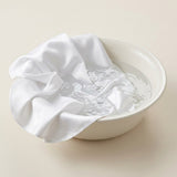 A white satin or silk fabric cloth draped over a white ceramic bowl containing water, set against a solid light beige background.
