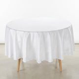 A round white tablecloth draped over a round table with light wood legs, set against a plain white wall and light gray floor.