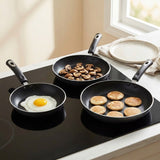 Three black nonstick frying pans with dark gray handles are arranged on a black glass stovetop in a bright kitchen. The pans contain a fried egg, sliced mushrooms, and small pancakes.