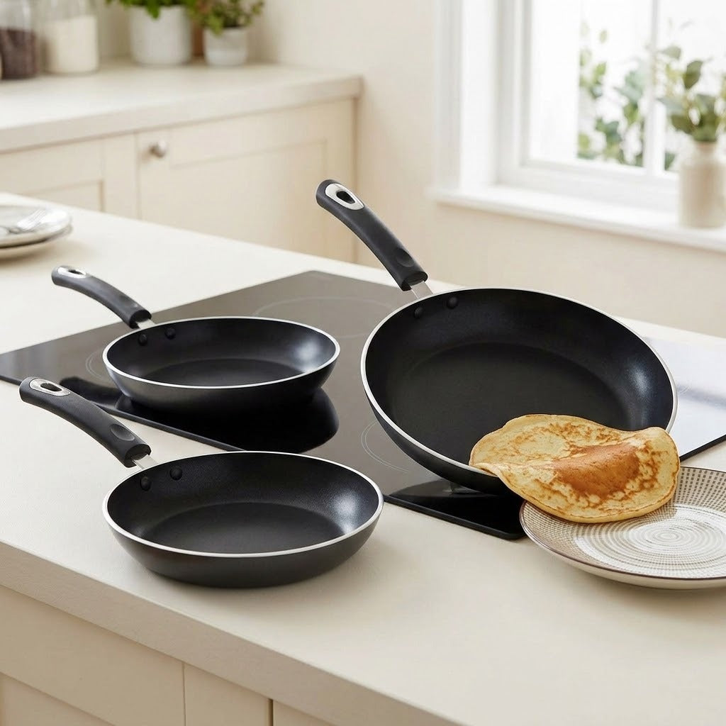 Three black nonstick frying pans with black handles are arranged on a white kitchen countertop beside a glass cooktop. A folded pancake sits on a plate in front of the largest pan.