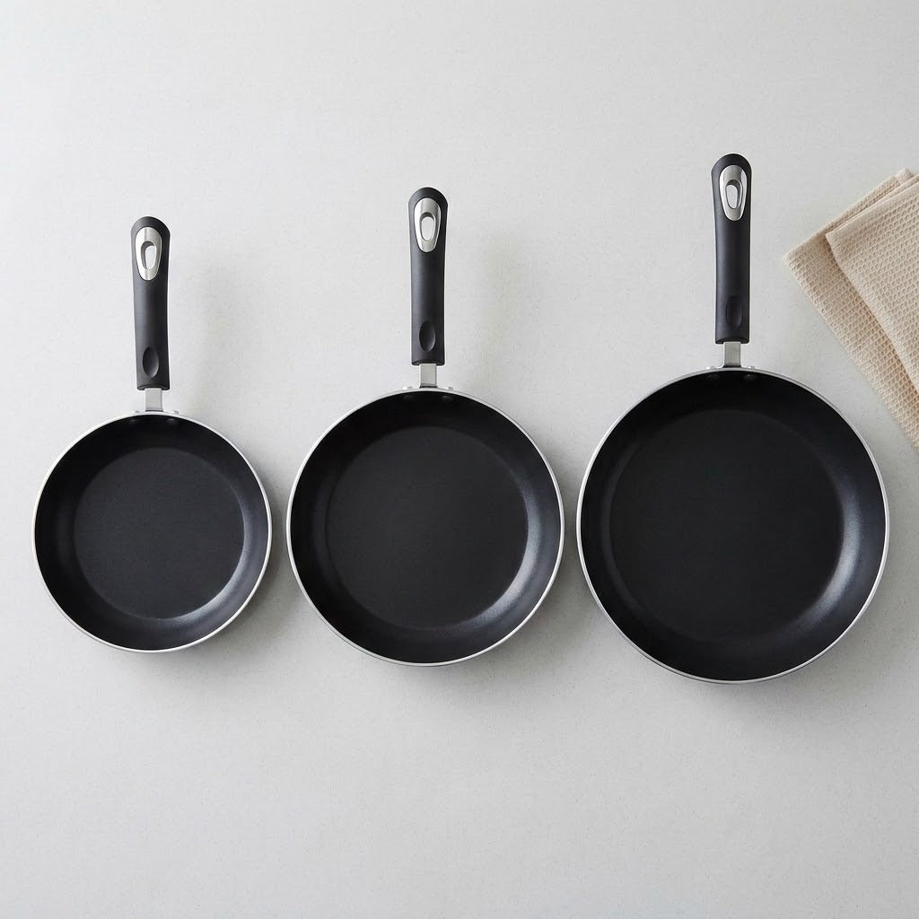 Three black non-stick frying pans of different sizes hanging on a light gray wall, with a beige cloth partially visible on the right side.