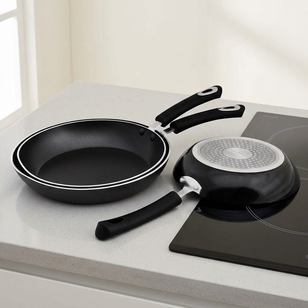 A black non-stick frying pan and a matching small saucepan with black handles and silver-rimmed edges, displayed on a light gray kitchen countertop beside a black induction cooktop.