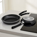 A black non-stick frying pan and a matching small saucepan with black handles and silver-rimmed edges, displayed on a light gray kitchen countertop beside a black induction cooktop.