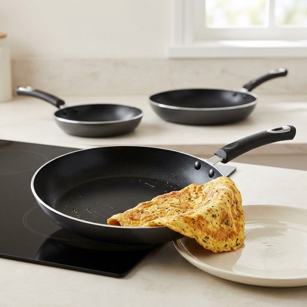 Three black nonstick frying pans with black handles are arranged on a light beige kitchen countertop. The front pan sits on a black induction cooktop with a folded omelet resting inside it, and a white plate is partially visible in the foreground.