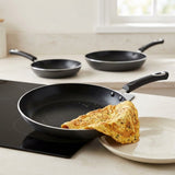 Three black nonstick frying pans with black handles are arranged on a light beige kitchen countertop. The front pan sits on a black induction cooktop with a folded omelet resting inside it, and a white plate is partially visible in the foreground.