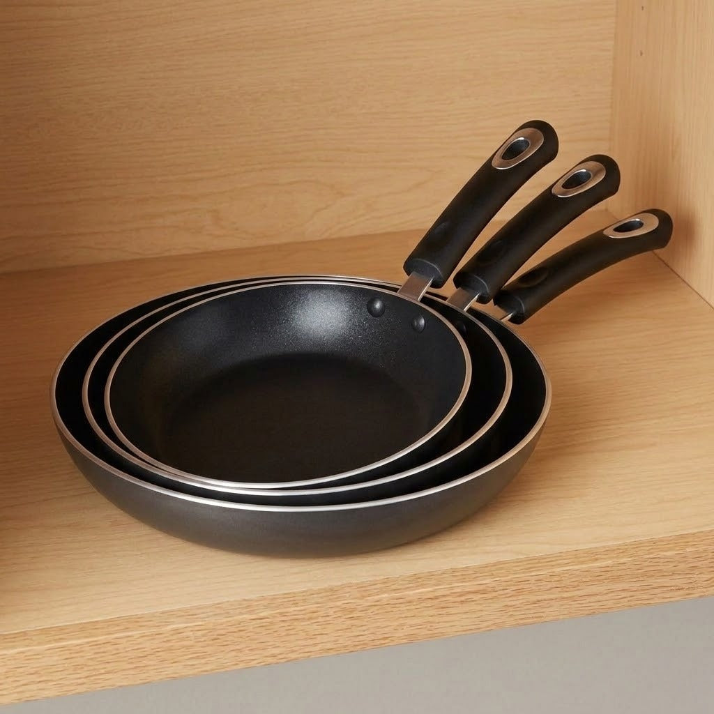 Three nested black non-stick frying pans with silver metal rims and black handles, stacked on a light wood shelf.