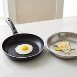 A black non-stick frying pan with a handle sits on a light countertop, cooking a sunny-side-up egg. A stainless steel pan is beside it with partially cooked egg pieces.