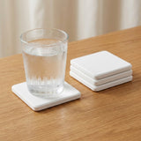 A set of four square white coasters on a light wood table, with one coaster holding a clear glass of water and three stacked coasters beside it.