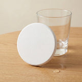 A round white coaster with a textured surface, shown next to a clear glass on a light wood tabletop.