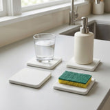 Four square white soap dish coasters arranged on a light countertop in a kitchen setting, including one holding a glass of water, one holding a cream-colored pump bottle with a silver dispenser, one empty, and one holding a green and yellow sponge.