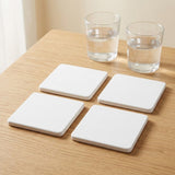 Four square white coasters arranged on a light wood table, with two clear drinking glasses in the background.