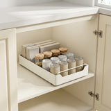 A white under-cabinet pull-out spice rack organizer shown inside an open kitchen cabinet, holding jars, boxes, and containers.