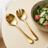 Two gold-colored salad servers, one spoon and one fork, arranged on a round cream-colored table next to a bowl of salad with greens, tomatoes, and cheese shavings.