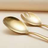 A close-up of two polished gold-toned utensils, one spoon and one fork, resting on a warm beige surface with a softly blurred background.