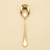 A single polished gold spoon with a reflective finish, centered vertically against a solid, warm cream-colored background.
