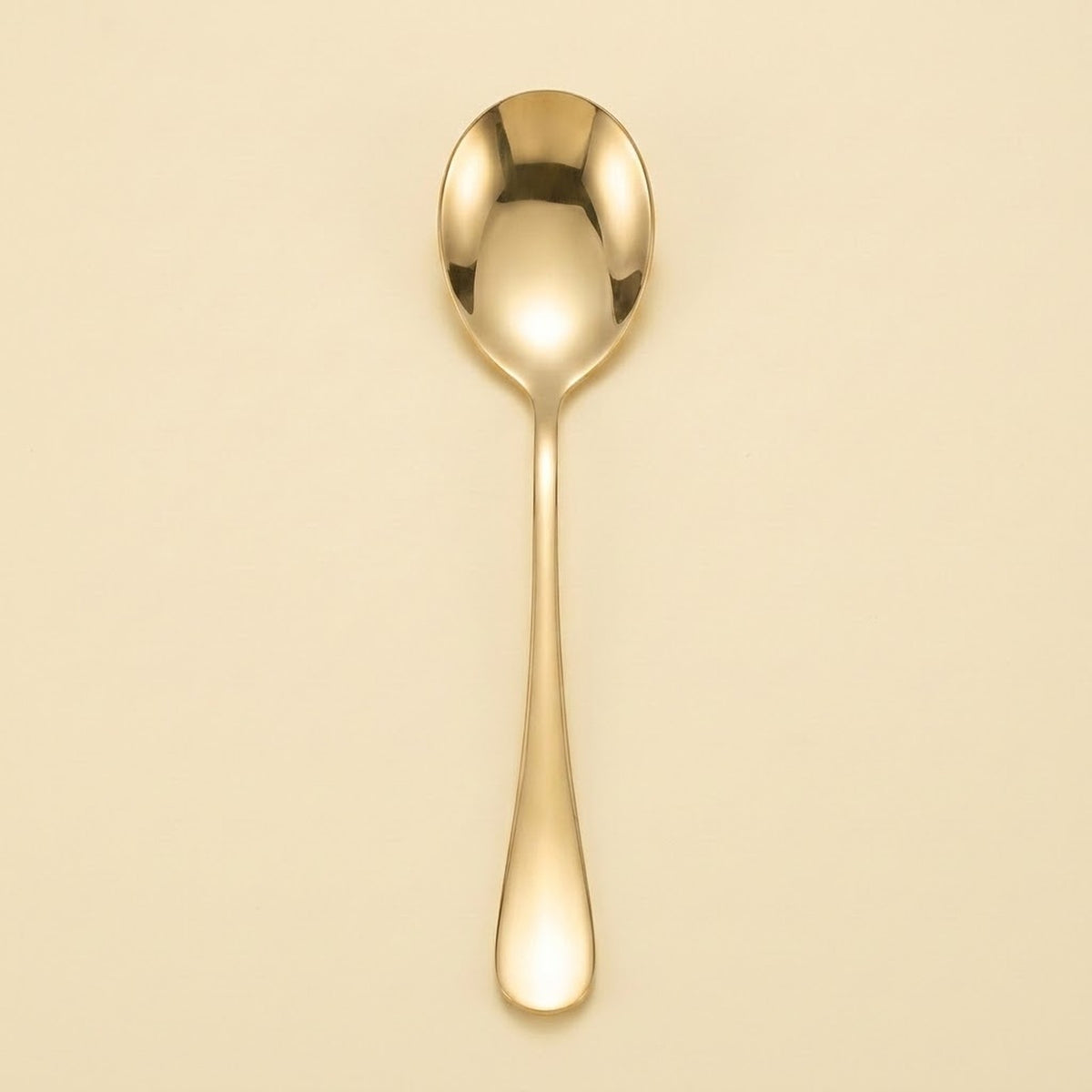 A single polished gold spoon with a reflective finish, centered vertically against a solid, warm cream-colored background.