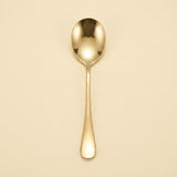 A single polished gold spoon with a reflective finish, centered vertically against a solid, warm cream-colored background.