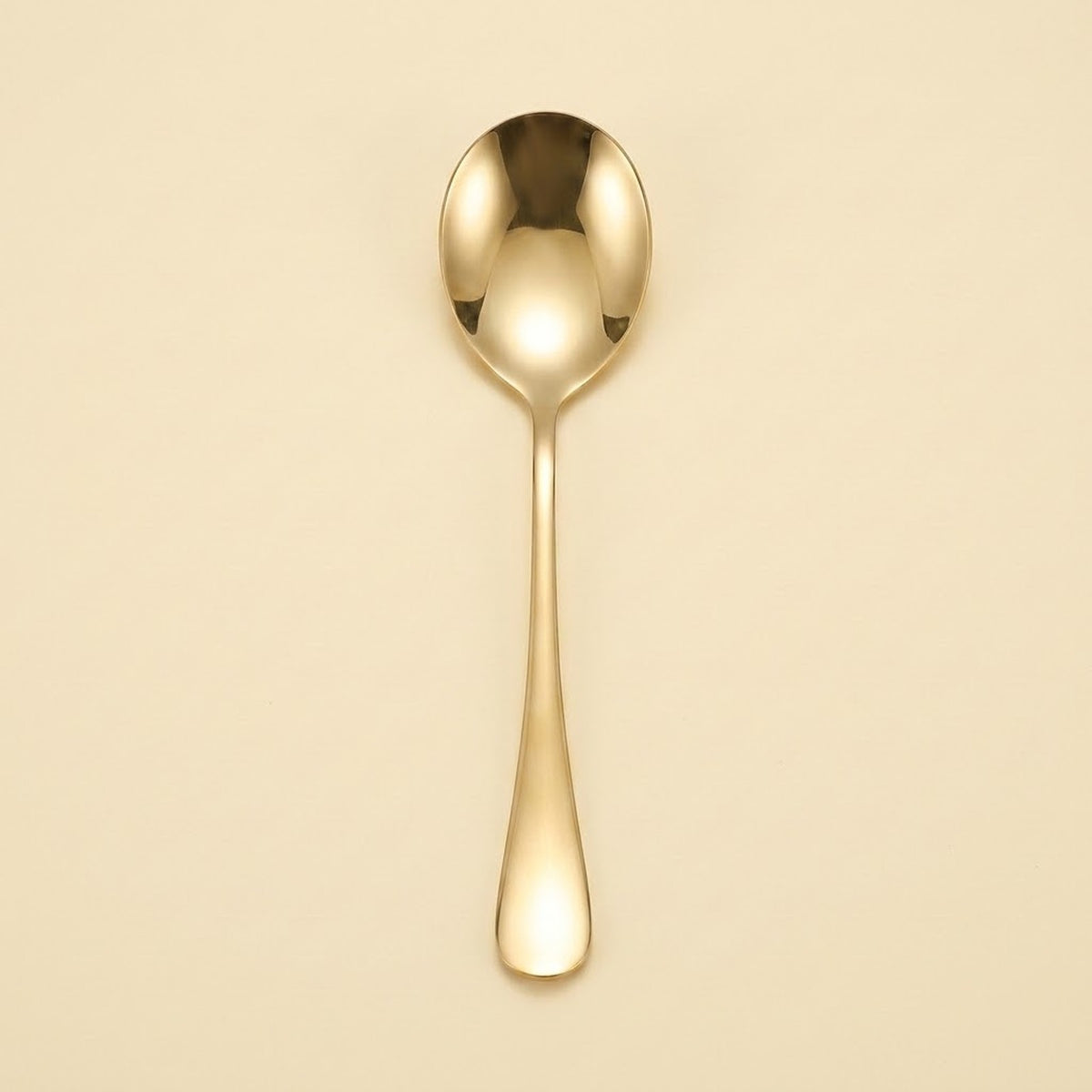 A single polished gold spoon with a reflective finish, centered vertically against a solid, warm cream-colored background.