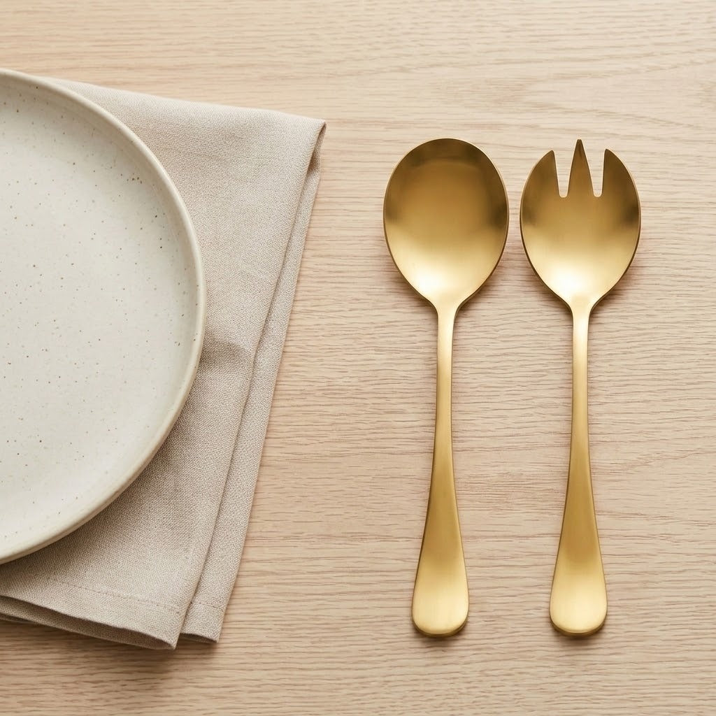 A gold-colored metal spoon and matching fork with a wide bowl, displayed on a light wood grain surface next to a folded beige linen napkin and a partial view of a speckled ceramic plate.