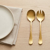A gold-colored metal spoon and matching fork with a wide bowl, displayed on a light wood grain surface next to a folded beige linen napkin and a partial view of a speckled ceramic plate.