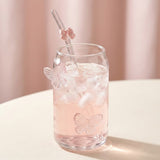 A clear glass can-shaped tumbler filled with a pink drink and ice, with butterfly-shaped glass accents and a clear straw decorated with a small pink bow, set on a light surface against a soft pink background.