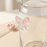 A clear glass jar with a translucent pink butterfly decoration attached to the side, photographed on a soft cream background.