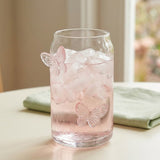 A clear glass can-shaped tumbler filled with ice and a pale pink drink, decorated with two translucent pink butterfly motifs, set on a tabletop with a folded green napkin in a softly blurred indoor setting.
