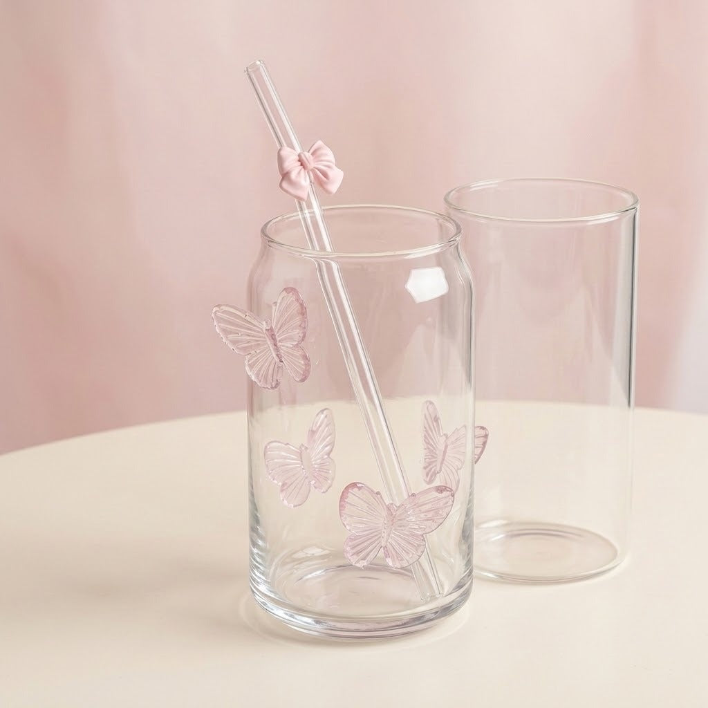 Clear glass can-shaped cup with pink butterfly decals and a clear straw with a pink bow, shown beside a second empty cup on a light beige surface against a soft pink background.