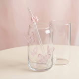 Clear glass can-shaped cup with pink butterfly decals and a clear straw with a pink bow, shown beside a second empty cup on a light beige surface against a soft pink background.