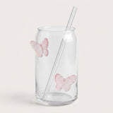Clear glass can-shaped tumbler with two pink butterfly decals and a clear glass straw, shown on a light beige background.