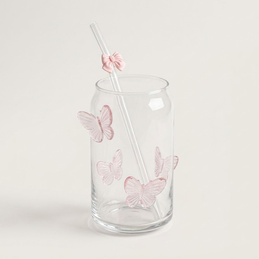 A clear glass tumbler with pink butterfly decorations and a clear straw featuring a pink bow accent, set against a plain light beige background.