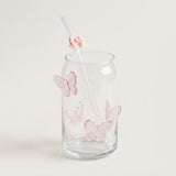 A clear glass tumbler with pink butterfly decorations and a clear straw featuring a pink bow accent, set against a plain light beige background.
