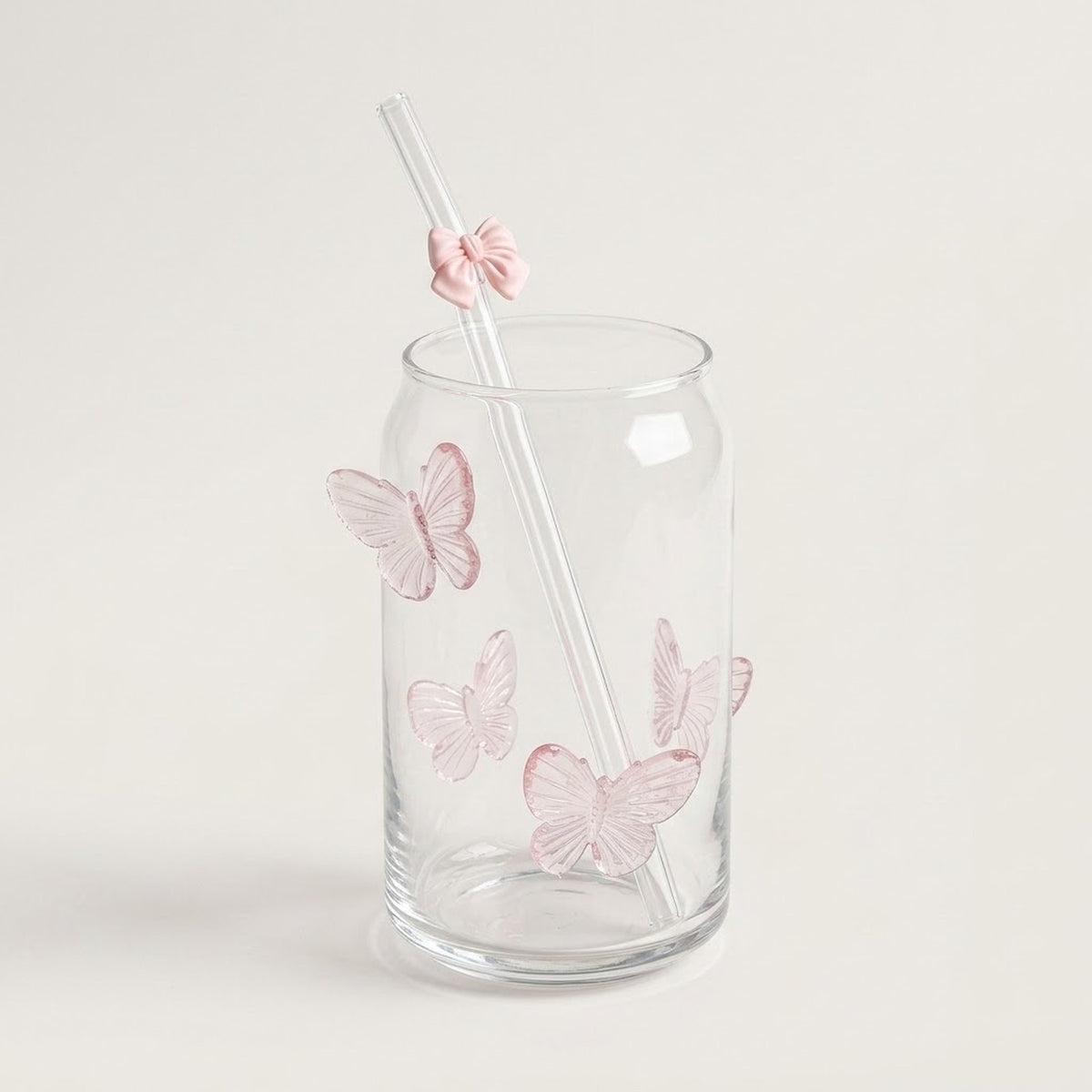 A clear glass tumbler with pink butterfly decorations and a clear straw featuring a pink bow accent, set against a plain light beige background.