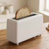 A white four-slot toaster with a slice of bread in the top slot, sitting on a wooden tabletop in a softly lit kitchen setting.