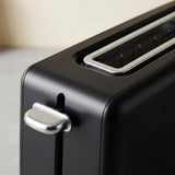 Close-up of a matte black toaster with a silver lever handle and metallic trim, shown against a softly blurred light beige background.