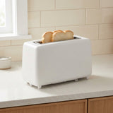 A white two-slot toaster sits on a light countertop with two slices of toast emerging from the top, set against a tiled kitchen backsplash.