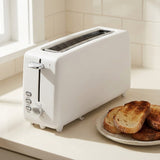 A white two-slot toaster sits on a light countertop in a bright kitchen setting, with slices of toasted bread on a plate nearby.