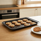 A black rectangular baking sheet with rounded corners holding several chocolate chip cookies, set on a wooden kitchen countertop with a white plate and a cookie nearby.