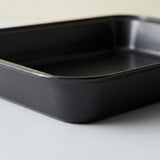 A black rectangular baking pan with rounded corners, photographed on a light gray surface against a neutral off-white background.