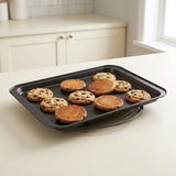 A dark gray rectangular baking sheet holding several chocolate chip cookies, set on a light countertop in a bright kitchen with white cabinets and a window in the background.