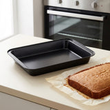 A rectangular black non-stick baking pan sits on a white kitchen countertop next to a baked loaf of cake on parchment paper, with a blurred oven in the background.