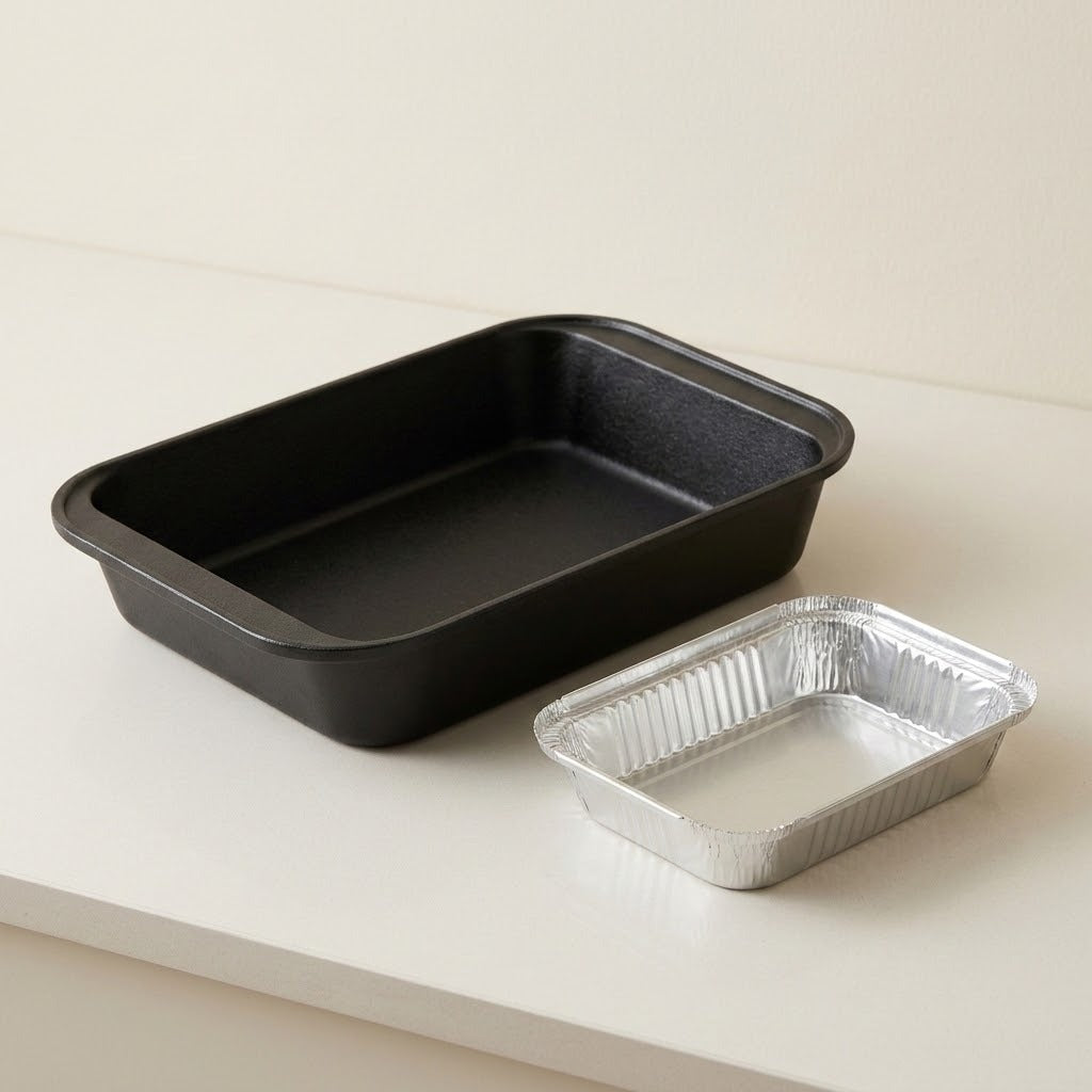 A rectangular black non-stick baking pan next to a smaller rectangular aluminum foil tray on a clean white surface.