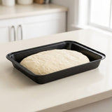 A black rectangular baking pan containing risen dough, set on a light beige countertop in a bright kitchen.